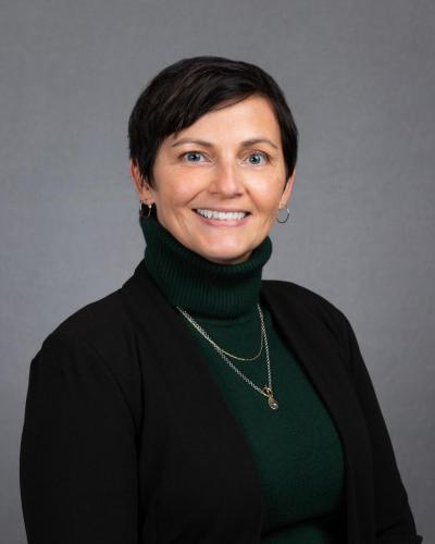 A smiling woman wearing a black blazer, green turtleneck, and gold jewelry against a gray background.