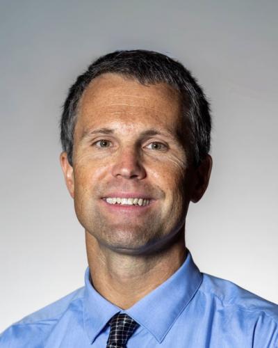 A man in a blue shirt and tie smiles at the camera against a gray background.