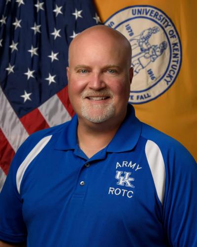 A man smiles while wearing a University of Kentucky ROTC blue polo shirt in front of an American flag and University of Kentucky flag.